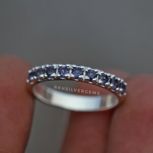 May include: A silver ring with a row of small, oval-shaped, purple gemstones. The ring is set with multiple stones, and the band is smooth and polished. The text "RKHSILVERGEMS" is engraved on the ring.
