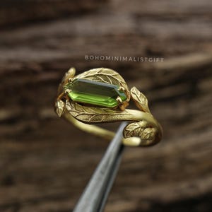 Könnte beinhalten: Ein goldfarbener Ring mit einem rechteckigen, im Baguetteschliff geschliffenen grünen Edelstein, der in eine Ringschiene in Form überlappender Blätter gefasst ist. Der Text BOHOMINIMALISTGIFT ist über dem Ring sichtbar. Die Schiene ist strukturiert.