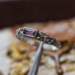 May include: A silver ring featuring a rectangular, purple, baguette-cut gemstone set in the center. The band is crafted with an intricate, raised relief pattern of small leaves and vines wrapping around the entire circumference, with the text R K H S I L V E R G E M S visible.