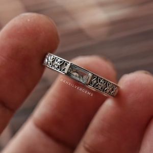 May include: A silver ring featuring a rectangular, clear stone set in the center, flanked by intricate, repeating knotwork patterns along the band. The text RKHSILVERGEMS is engraved on the side of the band near the stone setting.