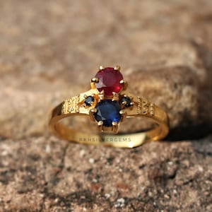 May include: A gold ring featuring a round red gemstone at the top, a round blue gemstone below it, and two small black accent stones on the sides. The band has a textured, patterned design on both sides. The text RKHSILVERGEMS is visible.