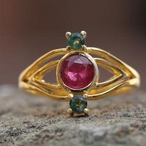 Peut inclure: Une bague en or ornée d'un rubis central et de deux émeraudes. La bague présente un design unique avec une monture fendue et des courbes décoratives. La bague est posée sur une surface en pierre grise.