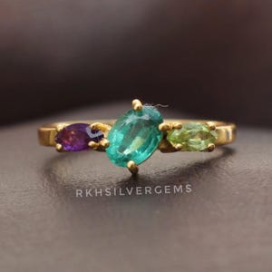 Peut inclure: Une bague en or ornée de trois pierres précieuses : une pierre centrale ovale de couleur teal, flanquée d'une pierre violette et d'une pierre verte. La bague est posée sur un fond sombre. Le texte "RKHSILVERGEMS" est visible.