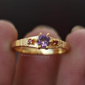Peut inclure: Une bague en or ornée d'une pierre centrale violette et de deux petites pierres rouges. La monture présente un motif texturé. Le texte "RKHSILVERGEMS" est visible sur la bague.