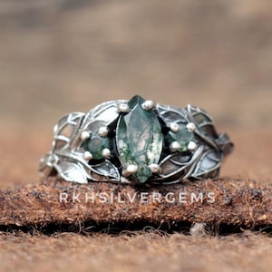 May include: A silver ring featuring a marquise-cut moss agate gemstone, flanked by smaller round gemstones. The ring has a leaf design on the band and around the stones. The text "RKHSILVERGEMS" is visible on the surface.