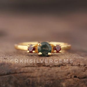 Könnte beinhalten: Ein goldener Ring mit einem zentralen dunkelgrünen Edelstein, flankiert von zwei kleineren roten Edelsteinen. Der Ring liegt auf einer Holzoberfläche, mit dem Text "RKHSILVERGEMS" sichtbar.