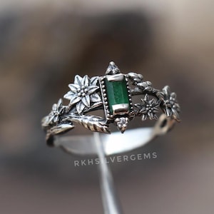 Può includere: Un anello d'argento con una gemma verde rettangolare al centro, circondato da motivi floreali e fogliari. L'anello ha un'estetica vintage o antica, con dettagli intricati e una finitura lucida. L'anello è tenuto da uno strumento.
