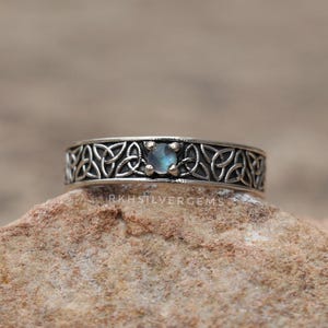 May include: A silver ring with a Celtic knot design. The ring features a small, round, light blue gemstone set in the center. The band is intricately detailed with the knot pattern. The ring is displayed on a textured, brown surface.