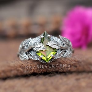 Puede incluir: Un anillo plateado con una piedra preciosa verde claro en el centro. El anillo está adornado con un detallado diseño de hojas. El texto "RKHSILVERGEMS" es visible en la parte inferior de la imagen. El anillo está sobre una superficie marrón.