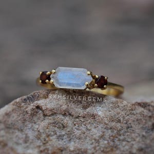 May include: A gold ring featuring a central horizontal hexagonal moonstone flanked by two small round dark red garnet stones. The band is thin and metallic gold. The text RKHSILVERGEMS is visible on the stone setting. The ring is resting on a rock.