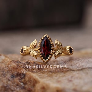 May include: A gold ring featuring a central marquise-cut dark red gemstone surrounded by a beaded border, flanked by leaf-shaped metal accents and smaller round red stones on each side. The text RKHSILVERGEMS is printed below the ring.