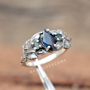 May include: A silver ring with a dark blue, pear-cut gemstone at its centre, complemented by smaller green stones. The ring's band is decorated with leaf patterns. The text "RKHILVERGEMS" is visible.