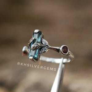 May include: A silver ring featuring a rectangular, light blue gemstone as the focal point. The ring has a nature-inspired design with leaf accents and a small, round, red gemstone. The text "RKHSILVERGEMS" is visible.