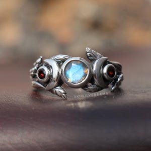 Peut inclure: Bague en argent ornée d'une pierre bleue centrale et de deux pierres rouges, serties dans un motif floral et de feuilles. Le texte "RKHSILVERGEMS" est visible. La bague est présentée sur une surface marron.