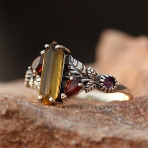 Könnte beinhalten: Ein silberner Ring mit einem großen, rechteckigen gelbbraunen Edelstein als Mittelpunkt. Zwei kleinere rote Edelsteine und ein violetter Edelstein akzentuieren das Design, zusammen mit Blatt- und Blumendetails. Der Ring liegt auf einer strukturierten, braunen Oberfläche.