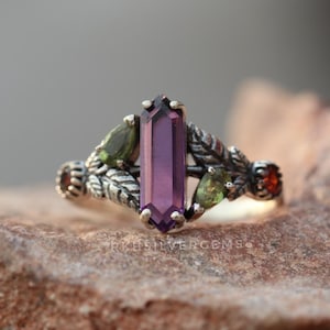 May include: A silver ring with a central, rectangular purple gemstone. The ring is decorated with green and red gemstones, and leaf motifs. The ring is displayed on a natural stone surface.