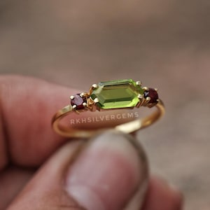 May include: A gold ring with a central, light green, emerald-cut gemstone and two smaller, round, dark red gemstones. The ring has a delicate band and is held in a hand. The text "RKHSILVERGEMS" is visible on the ring.
