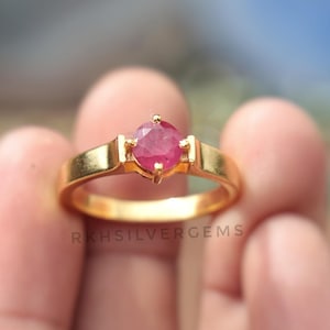 May include: A gold ring with a bright red ruby gemstone. The ring has a plain band with rectangular details either side of the ruby. The text "RKHSILVERGEMS" is visible on the ring.