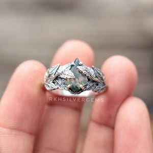 Puede incluir: Un anillo de plata con una piedra preciosa de ágata musgosa en forma de pera. El anillo está rodeado por un diseño de hojas detallado. El texto "RKHSILVERGEMS" es visible en el anillo.