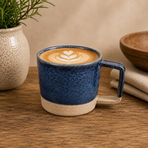 May include: A ceramic mug with a speckled dark blue glazed upper section and a matte beige base. The mug features a square-shaped handle and is filled with a coffee beverage topped with white foam arranged in a symmetrical leaf-like latte art pattern.