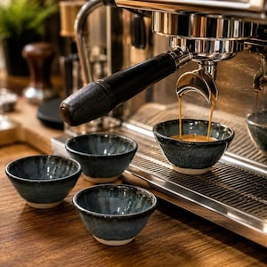 May include: Three small, round, dark blue ceramic bowls with a speckled glaze and a light-coloured base. One bowl is positioned directly under the metal portafilter of an espresso machine, catching a stream of freshly brewed dark brown coffee.