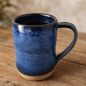 May include: A ceramic mug featuring a deep blue speckled glaze with a dark rim and a handle. The base of the mug is unglazed, revealing a natural tan clay texture. The cylindrical body has a slightly tapered shape with a smooth, glossy finish.