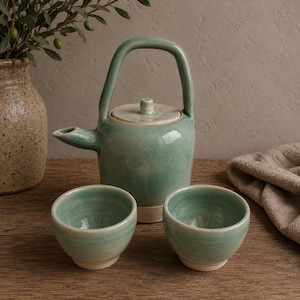 May include: A celadon green ceramic tea set. The set comprises a teapot with a curved handle, a lid, and two small cups. The teapot and cups have a white base. The set is displayed on a wooden surface.