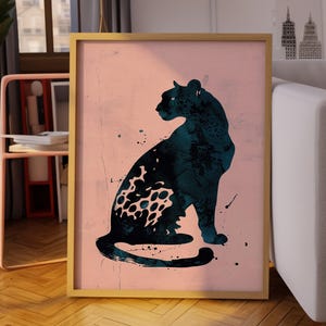 May include: A rectangular framed wall art print featuring a dark teal silhouette of a seated leopard against a solid light pink background. The leopard design incorporates a textured, ink-wash style with visible splatter details and a spotted pattern.