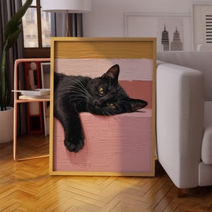 May include: A framed artwork featuring a black cat with yellow eyes resting on a pink and cream-colored background. The artwork is in a gold frame and is placed in a living room setting with a sofa and window.