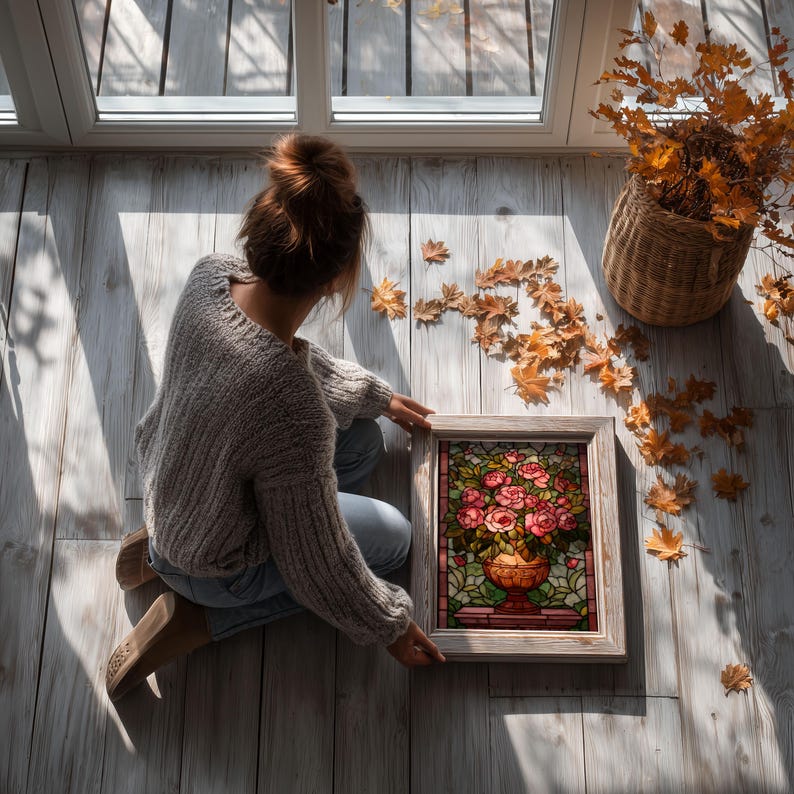 May include: A framed stained glass artwork featuring a bouquet of pink roses in a decorative vase. The artwork is set against a backdrop of green and red hues, framed in a light-colored wooden frame. The scene is set on a wooden floor with autumn leaves.