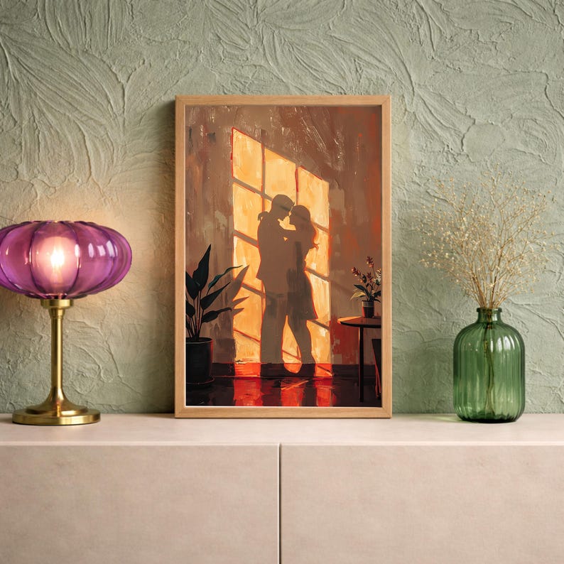 May include: A framed artwork depicts a couple embracing in front of a sunlit window. The warm color palette includes shades of orange, brown, and gold. A purple lamp and a green vase with dried flowers are on a white surface.