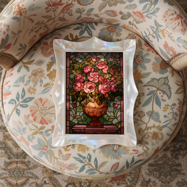 May include: A framed stained glass artwork featuring a bouquet of pink roses in a decorative vase. The frame is white with an ornate, scalloped edge. The artwork is set against a floral patterned chair.