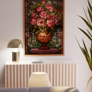 May include: A framed stained glass panel featuring a vase overflowing with pink roses and green leaves. The artwork is set against a backdrop of green and brown glass, framed in a light wood. The panel is displayed above a white cabinet.