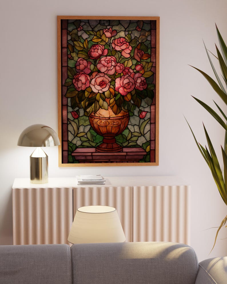 May include: A framed stained glass panel featuring a vase overflowing with pink roses and green leaves. The artwork is set against a backdrop of green and brown glass, framed in a light wood. The panel is displayed above a white cabinet.