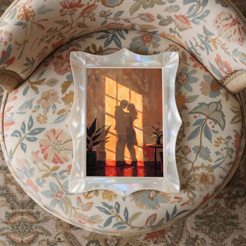 May include: A framed artwork featuring a couple embracing in front of a window, bathed in warm sunlight. The frame is white with an ornate, scalloped design. The artwork is set on a floral patterned chair.