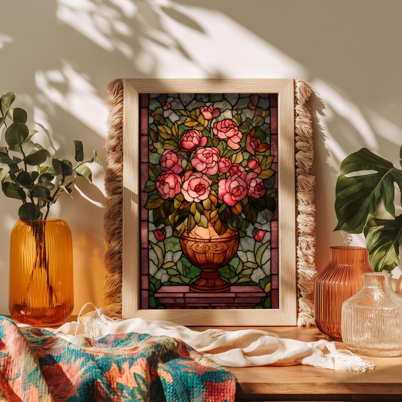 May include: A framed stained glass artwork featuring a bouquet of pink roses in a decorative vase. The artwork is set against a backdrop of green and pink glass, framed in light wood with a fringed edge. The artwork is displayed on a wooden surface.