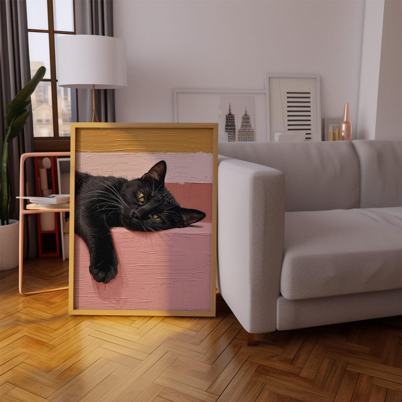 May include: A framed artwork featuring a black cat with yellow eyes resting on a pink and cream-colored background. The artwork is in a gold frame and is placed in a living room setting with a sofa and window.