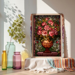 May include: A framed stained glass artwork featuring a bouquet of pink roses in a decorative vase. The artwork is set against a backdrop of green leaves and is framed with a light wood border. The frame is decorated with colorful fringe.