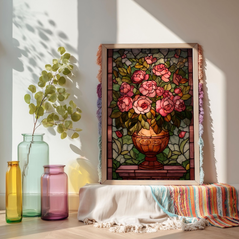 May include: A framed stained glass artwork featuring a bouquet of pink roses in a decorative vase. The artwork is set against a backdrop of green leaves and is framed with a light wood border. The frame is decorated with colorful fringe.