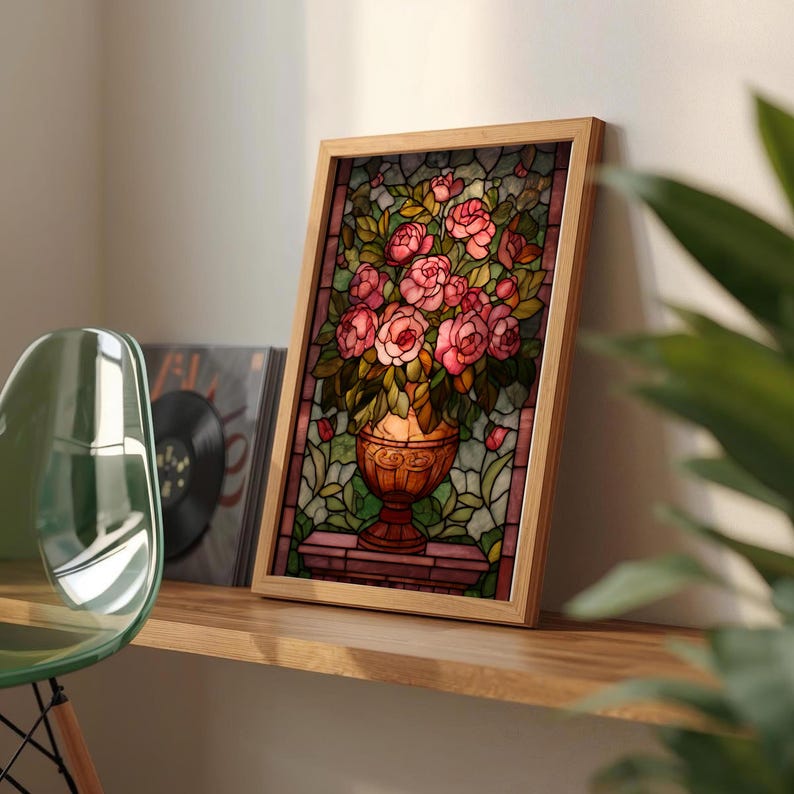May include: A framed stained glass artwork featuring a bouquet of pink roses in a decorative vase. The artwork is set against a backdrop of green foliage and is framed in light wood. The artwork is displayed on a wooden shelf.