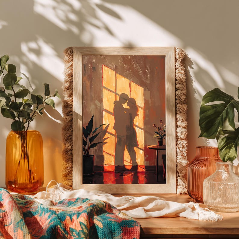 May include: A framed artwork depicts a couple embracing in front of a sunlit window. The painting has warm tones of orange and brown. The frame is light-colored wood with a fringed border. Decorative vases and plants are arranged around the artwork.