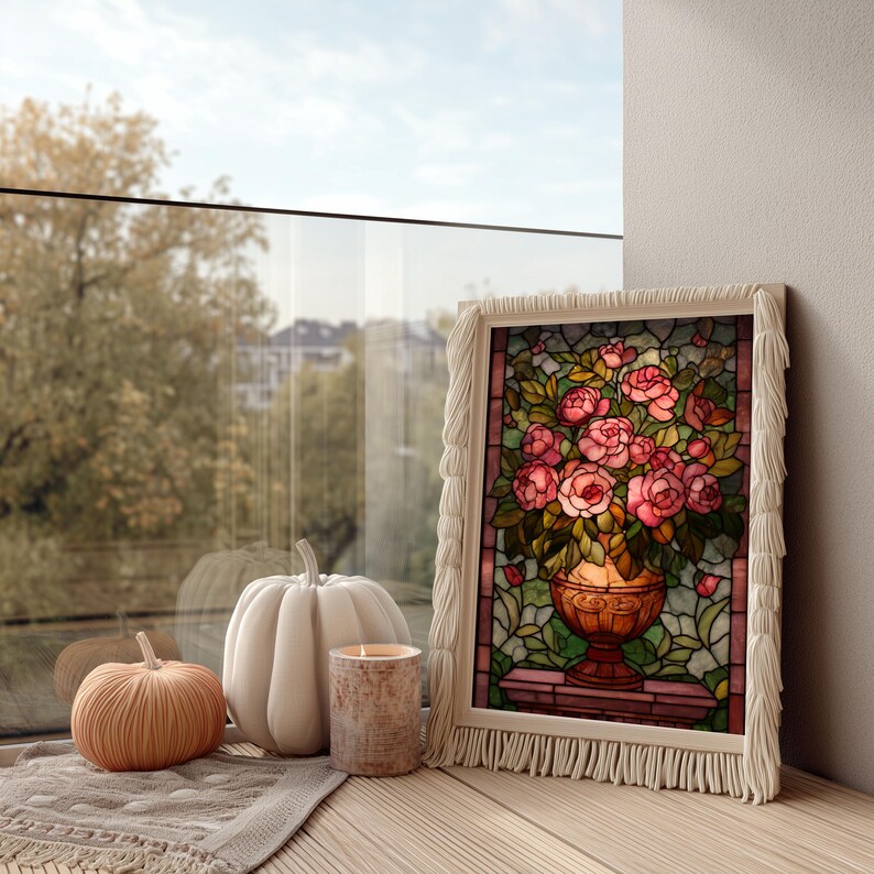 May include: A framed stained glass art piece featuring a bouquet of pink roses in a gold vase. The frame has a white fringe. Beside it are two pumpkins and a candle on a wooden surface.