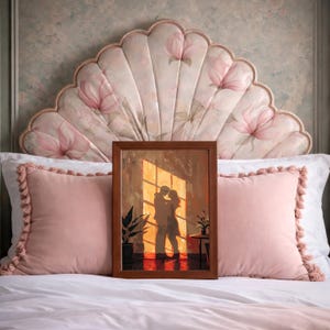 May include: A framed artwork depicts a couple embracing in front of a sunlit window. The artwork is in a brown frame and is propped up on a bed with pink pillows and a floral headboard. The scene is warm and inviting, with a focus on the couple's silhouette.