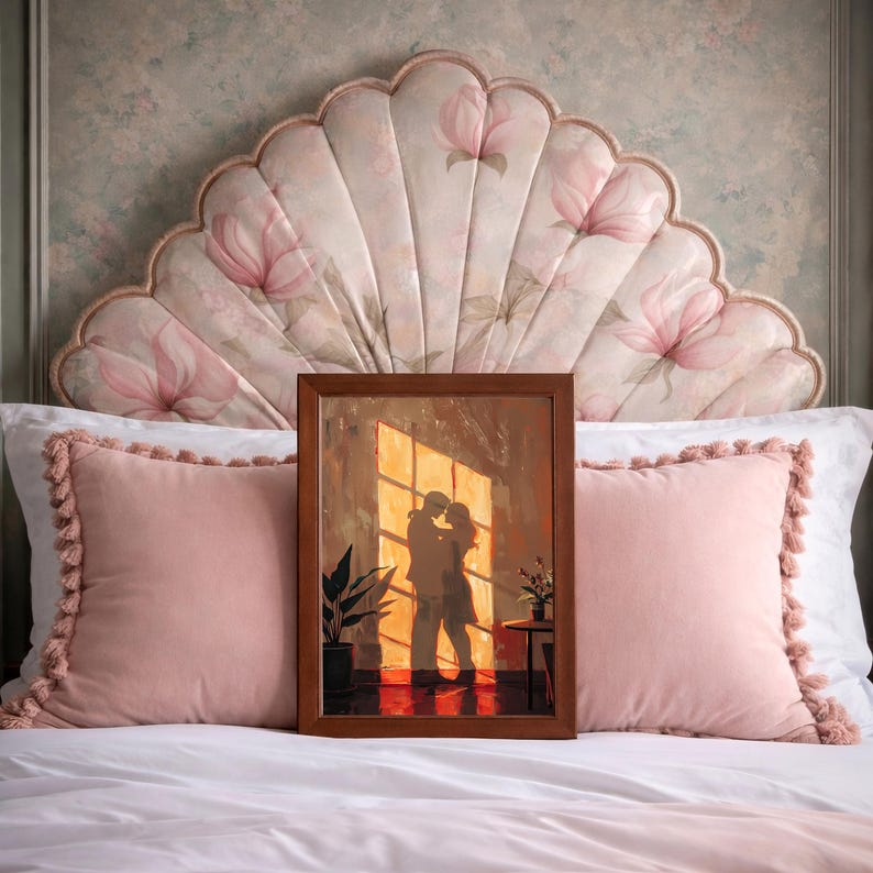 May include: A framed artwork depicts a couple embracing in front of a sunlit window. The artwork is in a brown frame and is propped up on a bed with pink pillows and a floral headboard. The scene is warm and inviting, with a focus on the couple's silhouette.