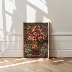 May include: A framed stained glass artwork featuring a bouquet of pink roses in a decorative vase. The artwork has a gold frame and is set against a background of green leaves and a brown border. The artwork is displayed on a wooden floor.