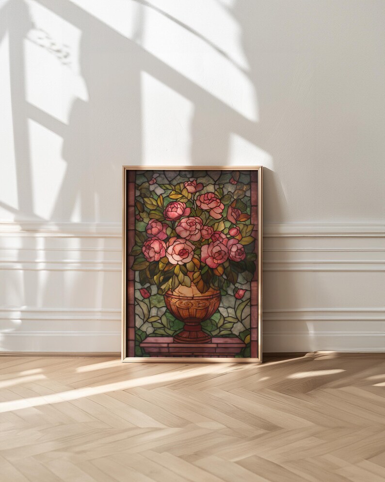 May include: A framed stained glass artwork featuring a bouquet of pink roses in a decorative vase. The artwork has a gold frame and is set against a background of green leaves and a brown border. The artwork is displayed on a wooden floor.