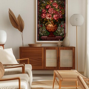 May include: A framed stained glass art piece featuring a bouquet of pink roses in a decorative vase. The artwork is set against a backdrop of green and brown stained glass, with a wooden frame. The image is displayed in a well-lit living room setting.