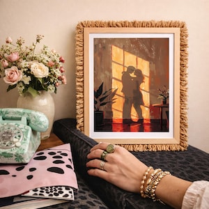 May include: A framed art print depicts a couple embracing in front of a sunlit window. The artwork is in a wooden frame with a fringed border. A vase of flowers, a vintage telephone, and a pink pouch with a black and white pattern are also visible.