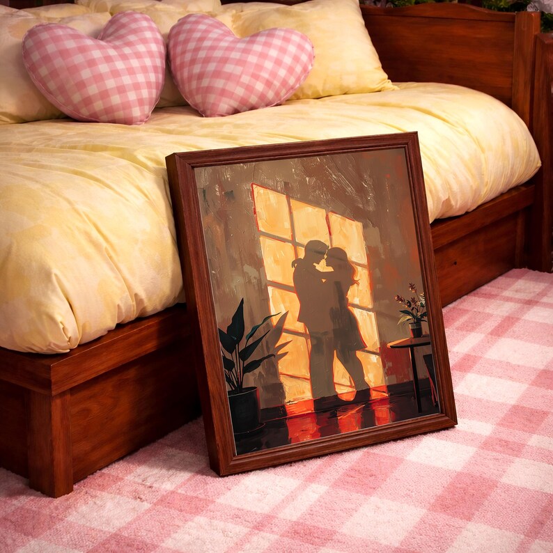 May include: A framed artwork depicting a couple embracing in front of a window, with warm sunlight streaming through. The artwork is in a brown frame, leaning against a bed with heart-shaped pillows and a pink and white checkered rug.