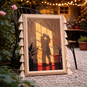 May include: A framed artwork depicting a couple embracing in front of a window, bathed in warm light. The artwork is in a wooden frame with decorative tassels. The scene includes a potted plant and a small table.